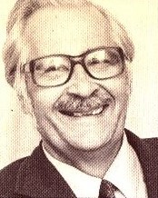 Erik Ewalds. Hymyilevä iäkäs mies, jolla on pyöreät silmälasit, puku ja solmio, vaalealla taustalla.
