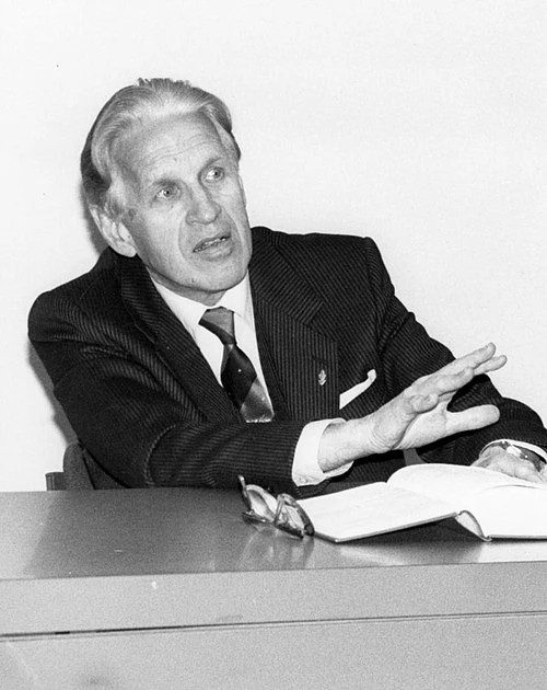 Paavo Hiltunen. Mies puvussa istuu pöydän ääressä ja elehtii käsillään, edessä avoin kirja.