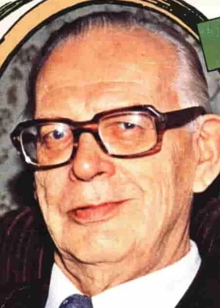 Tapio Nousiainen. Vanha mies mustissa kehyksissä ja sinisessä solmiossa, hymyilevä ilme, lähikuva.