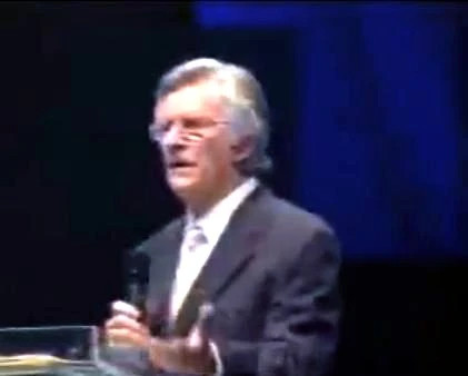 David Wilkerson. Harmaahiuksinen mies puhuu lavalla tummassa puvussa ja pitelee mikrofonia sinistä taustaa vasten.